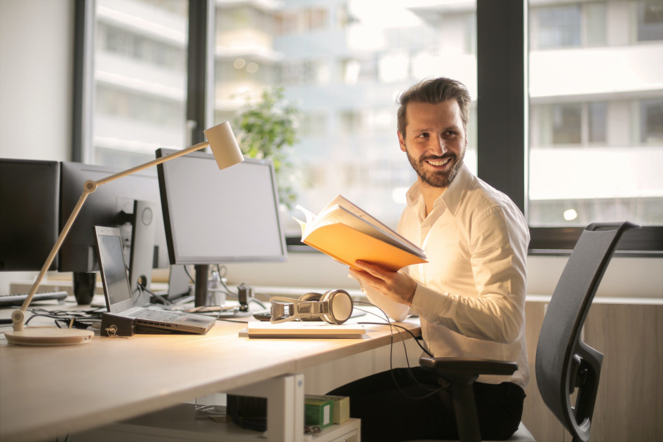 Foto de un oficinista sosteniendo unos folders, se mira sonriente sentado en su escritorio frente a un equipo de cómputo